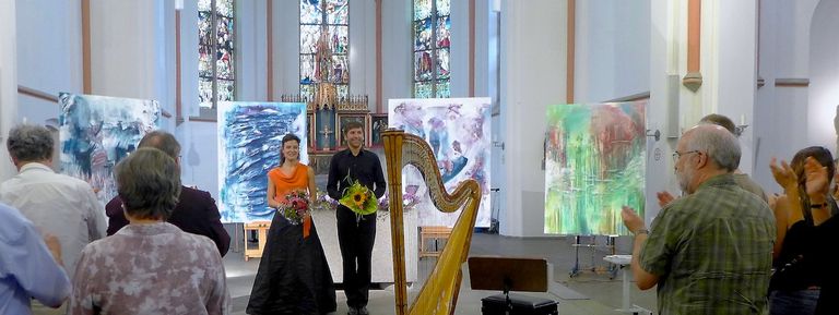 Applaus nach der Uraufführung von „Lieder in Bildern“. Das stehende Publikum beglückwünscht die beiden Musiker, die vor den großformatigen Bildern Ernst-Martin Heels stehen. Neben den Musikern steht die Harfe.
