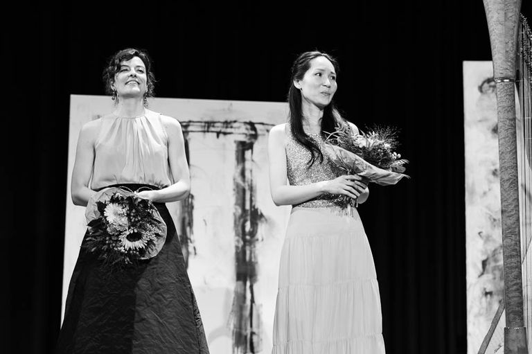 Zwei Musikerinnen nach einer Aufführung von „Lieder in Bildern“. Beide halten Blumen in der Hand. Rechts ist ein Teil einer Harfe zu sehen. Im Hintergrund stehen großformatige Gemälde von Ernst-Martin Heel